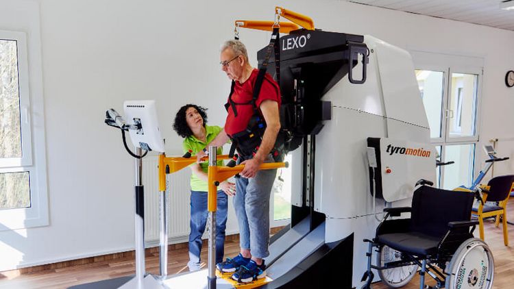 Ein Patient läuft mit Hilfe eines Gangtrainers, eine Therapeutin zeigt ihm, wie das funktioniert..